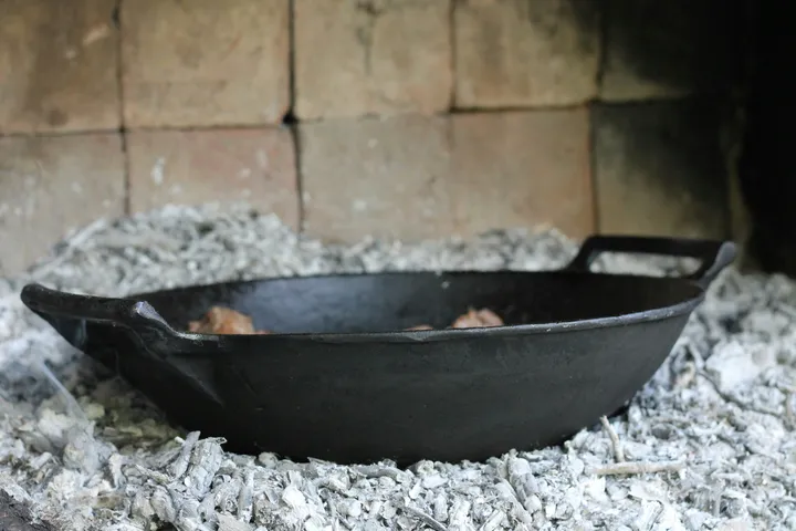 A vintage-style cast iron skillet resting in fireplace ash with worn dark seasoning, the look of decades of use