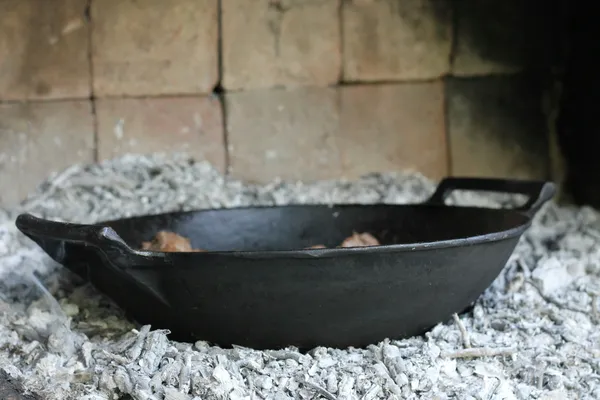 A vintage-style cast iron skillet resting in fireplace ash with worn dark seasoning, the look of decades of use