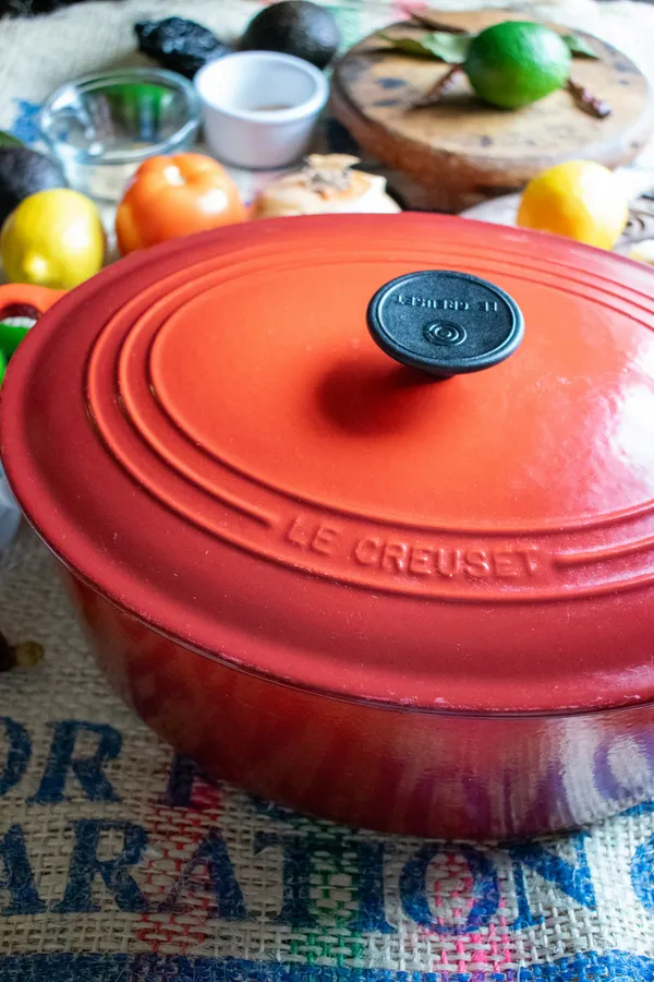 A red Le Creuset enameled cast iron dutch oven with the lid on, surrounded by fresh produce