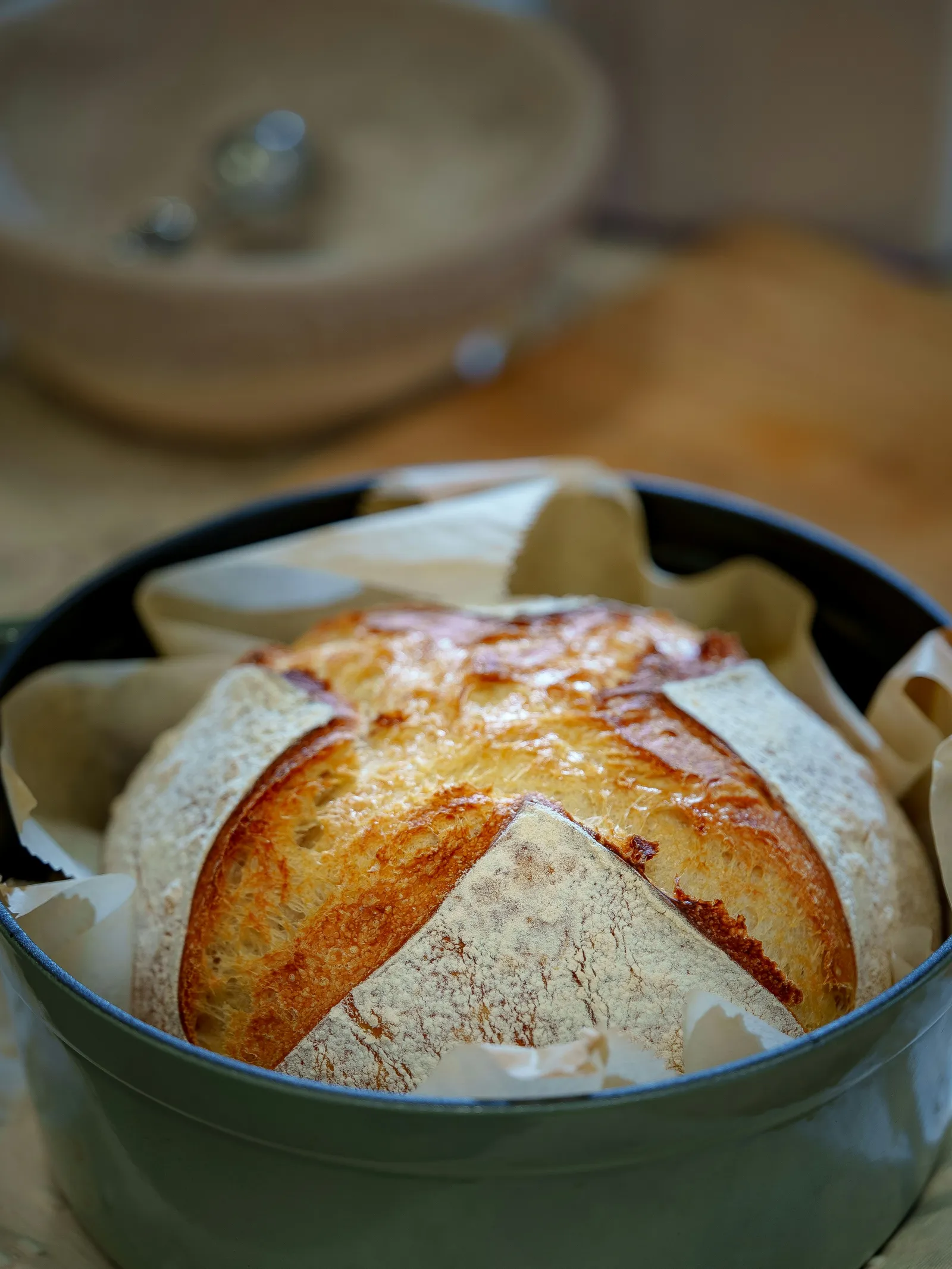 A golden cast iron dutch oven bread loaf with a scored cross, baked in a sage-green enameled dutch oven on parchment paper