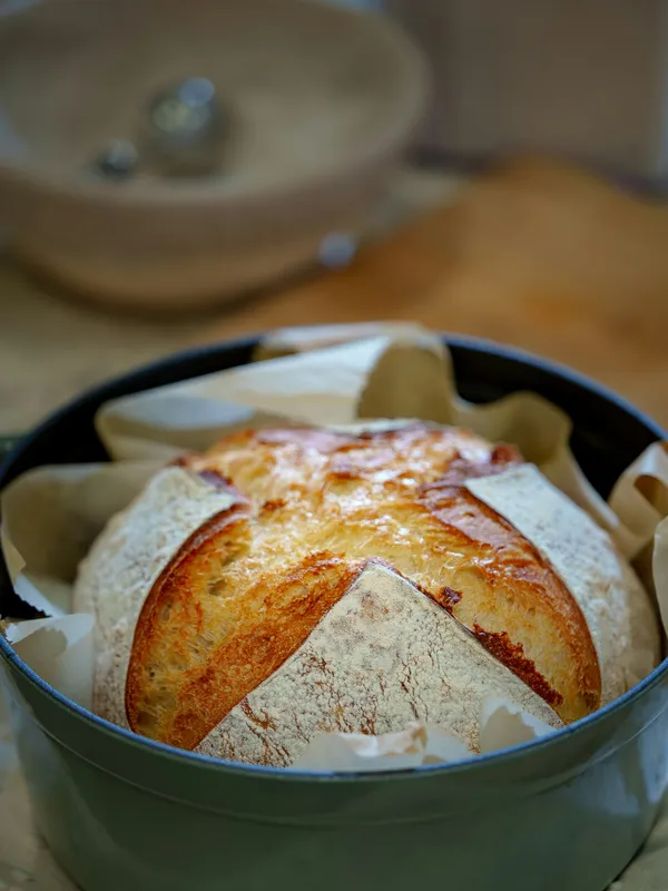 A golden cast iron dutch oven bread loaf with a scored cross, baked in a sage-green enameled dutch oven on parchment paper